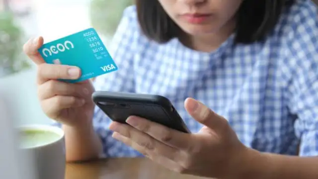 uma mulher mexendo no celular e com o cartão Neon Visa na outra mão